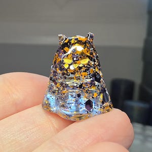 Op de afbeelding: Een klein, doorschijnend, geestvormig harsfiguur met een heldere basis die overgaat in een dichte mix van zwarte, oranje en gouden zeshoekige glitterdeeltjes door het bovenlichaam en de puntige oren heen.