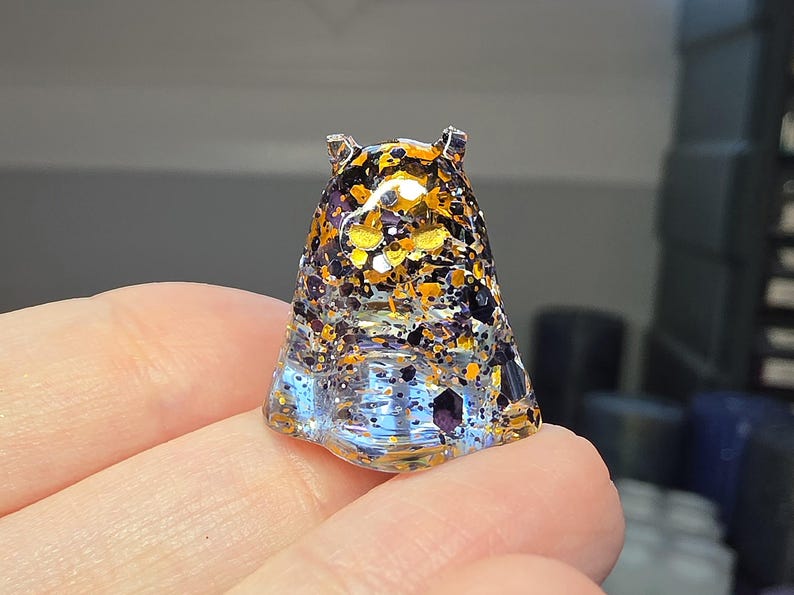 Op de afbeelding: Een klein, doorschijnend, geestvormig harsfiguur met een heldere basis die overgaat in een dichte mix van zwarte, oranje en gouden zeshoekige glitterdeeltjes door het bovenlichaam en de puntige oren heen.