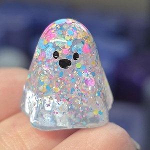 Op de afbeelding: Een klein, doorschijnend, spookvormig beeldje gemaakt van heldere hars gevuld met een dichte mix van veelkleurige, iriserende en holografische glitters in tinten roze, blauw, geel en zilver, voorzien van twee zwarte ogen en een zwarte mond.