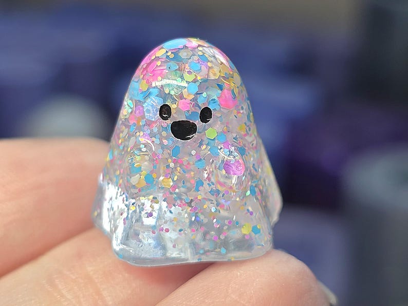 Op de afbeelding: Een klein, doorschijnend, spookvormig beeldje gemaakt van heldere hars gevuld met een dichte mix van veelkleurige, iriserende en holografische glitters in tinten roze, blauw, geel en zilver, voorzien van twee zwarte ogen en een zwarte mond.