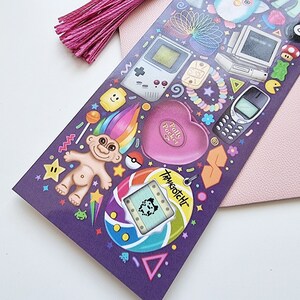 Pode incluir: Uma folha de adesivos roxa com &iacute;cones nost&aacute;lgicos dos anos 90, como um Game Boy, Polly Pocket e Tamagotchi. Uma borla rosa est&aacute; presa no canto superior esquerdo. O fundo &eacute; rosa claro.