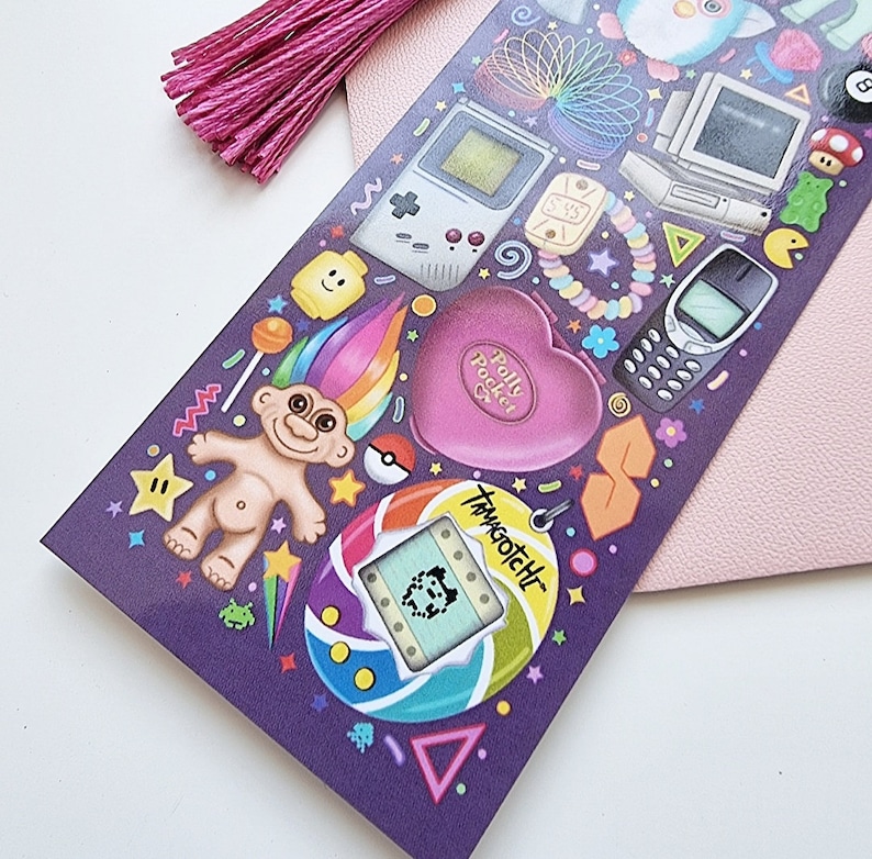 Pode incluir: Uma folha de adesivos roxa com &iacute;cones nost&aacute;lgicos dos anos 90, como um Game Boy, Polly Pocket e Tamagotchi. Uma borla rosa est&aacute; presa no canto superior esquerdo. O fundo &eacute; rosa claro.