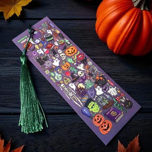 Può includere: Un segnalibro viola a tema Halloween con un nappa verde. Il segnalibro è ricoperto di illustrazioni colorate di zucche, fantasmi, scheletri e altre icone spettrali. Una zucca arancione e foglie autunnali sullo sfondo.
