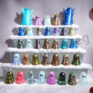 May include: A display of glittery ghost figurines in various colors, arranged on tiered platforms with faux snow. The ghosts have different patterns and colors, including blue, purple, gold, and red. A festive, decorative theme.