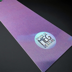 Puede incluir: Un marcap&aacute;ginas morado con un efecto brillante y una cuerda verde. El marcap&aacute;ginas presenta una pegatina circular con el texto "Scrambled MFG Designs" en una fuente estilizada. El fondo es negro.