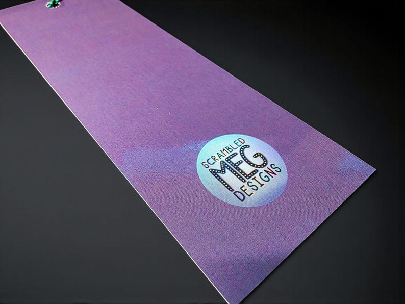 Puede incluir: Un marcap&aacute;ginas morado con un efecto brillante y una cuerda verde. El marcap&aacute;ginas presenta una pegatina circular con el texto "Scrambled MFG Designs" en una fuente estilizada. El fondo es negro.