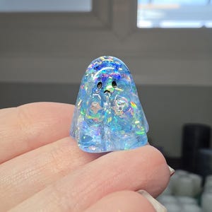 Op de afbeelding: Een klein, doorschijnend, lichtblauw spookfiguurtje gemaakt van hars. Het figuurtje heeft een ingebedde iriserende, holografische glitterafwerking die verschillende kleuren reflecteert. Het heeft twee eenvoudige zwarte stippen als ogen en een kleine zwarte mond.