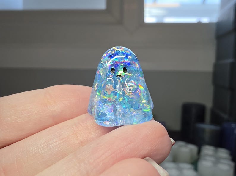 Op de afbeelding: Een klein, doorschijnend, lichtblauw spookfiguurtje gemaakt van hars. Het figuurtje heeft een ingebedde iriserende, holografische glitterafwerking die verschillende kleuren reflecteert. Het heeft twee eenvoudige zwarte stippen als ogen en een kleine zwarte mond.