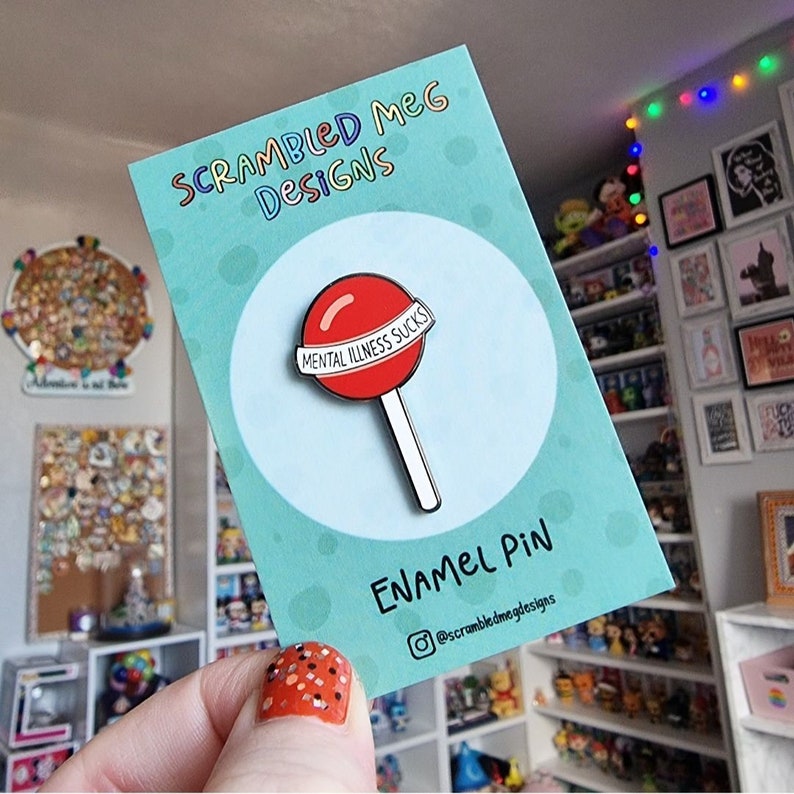 May include: Enamel pin featuring a red lollipop with the text "MENTAL ILLNESS SUCKS" on a white banner. The pin is on a teal card with the text "SCRAMBLED MEG DESIGNS" and "ENAMEL PIN". The Instagram handle @scrambledmegdesigns is also visible.