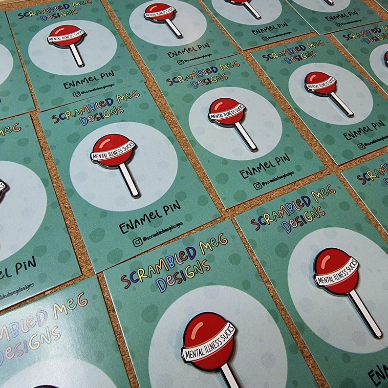 May include: Enamel pins featuring a red lollipop design with the text "Mental Illness Sucks" on a white banner. The pins are displayed on teal cards with the text "Scrambled Meg Designs".