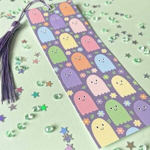 May include: A colorful bookmark featuring a pattern of smiling ghost characters in various pastel colors, set against a purple background with floral accents. The bookmark has a purple tassel attached to the top.