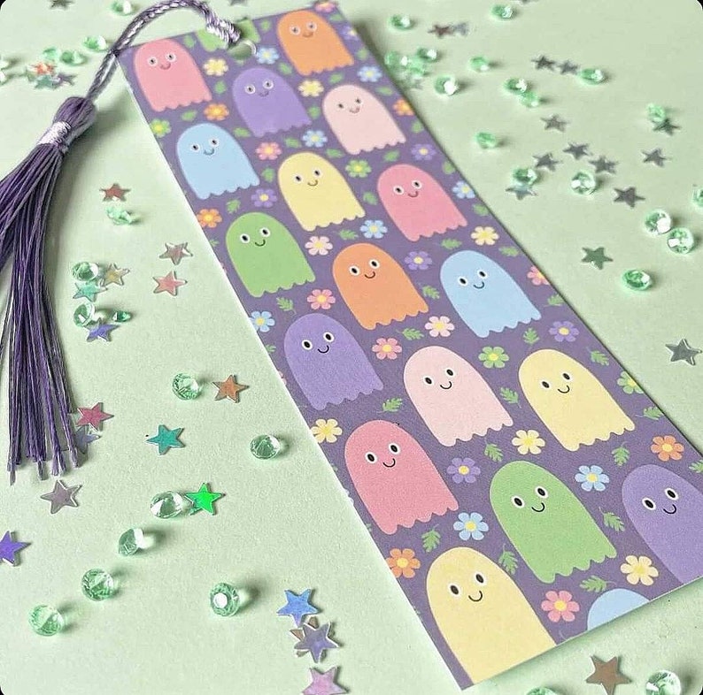 May include: A colorful bookmark featuring a pattern of smiling ghost characters in various pastel colors, set against a purple background with floral accents. The bookmark has a purple tassel attached to the top.