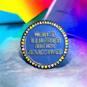 May include: A circular enamel pin featuring the text "MENTAL ILLNESSES ARE NOT ADJECTIVES" on a blue background. The pin has a decorative border with pastel colours. The background is a blur of rainbow hues.
