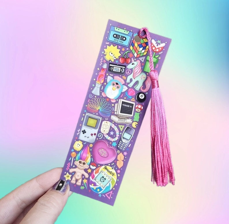 Pode incluir: Um marcador de livros roxo com ilustra&ccedil;&otilde;es dos anos 90, incluindo um Cubo de Rubik, Furby, Game Boy e um Tamagotchi. O marcador de livros tem uma borla rosa. O fundo &eacute; um gradiente de rosa, roxo e azul.