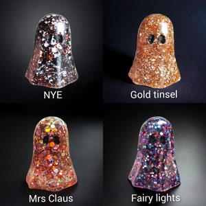 May include: Four glittery ghost figurines in various colors: black and silver, gold, orange and red, and purple with multi-colored glitter. Each ghost has two black eye holes. Text on the image reads "NYE", "Gold tinsel", "Mrs Claus", and "Fairy lights".