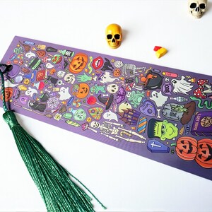 Puede incluir: Un marcap&aacute;ginas morado con una borla verde, con una ilustraci&oacute;n tem&aacute;tica de Halloween. El dise&ntilde;o incluye calabazas, fantasmas, esqueletos y otros elementos espeluznantes. Tambi&eacute;n hay dos peque&ntilde;as figuras de calaveras y caramelos.