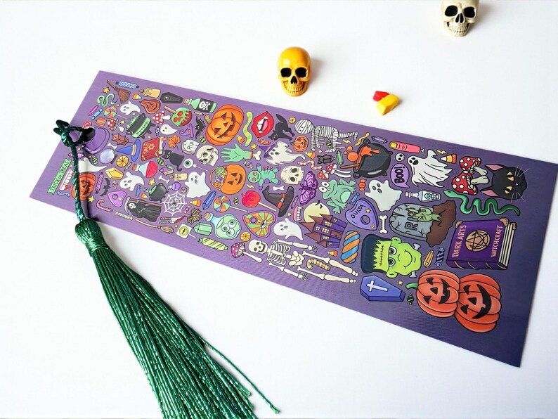 Puede incluir: Un marcap&aacute;ginas morado con una borla verde, con una ilustraci&oacute;n tem&aacute;tica de Halloween. El dise&ntilde;o incluye calabazas, fantasmas, esqueletos y otros elementos espeluznantes. Tambi&eacute;n hay dos peque&ntilde;as figuras de calaveras y caramelos.