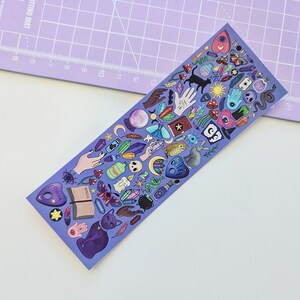 May include: A purple bookmark featuring a variety of mystical and occult-themed illustrations. The design includes a black cat, Ouija board, crystals, hands, mushrooms, and other magical symbols. The word "SPELLS" is visible on an open book.