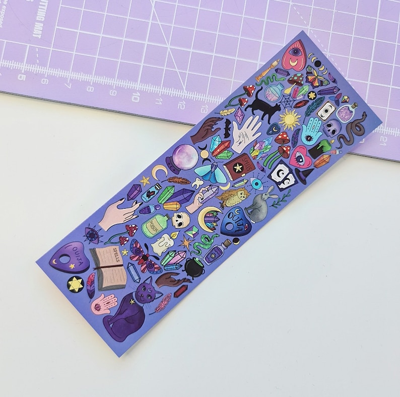 May include: A purple bookmark featuring a variety of mystical and occult-themed illustrations. The design includes a black cat, Ouija board, crystals, hands, mushrooms, and other magical symbols. The word "SPELLS" is visible on an open book.