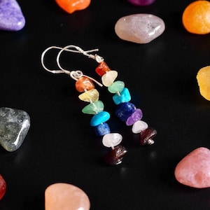 Peut inclure: Boucles d'oreilles en argent ornées de pierres précieuses colorées disposées en arc-en-ciel. Les boucles d'oreilles sont entourées de diverses pierres polies dans les tons de violet, orange, rose et vert, sur fond noir.
