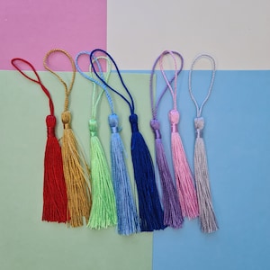 Pode incluir: Uma cole&ccedil;&atilde;o de borlas coloridas num arco-&iacute;ris de tons, incluindo vermelho, dourado, verde, azul, roxo, rosa e prateado. Cada borla apresenta um cord&atilde;o em la&ccedil;o para pendurar. As borlas est&atilde;o dispostas sobre um fundo rosa, verde e azul.