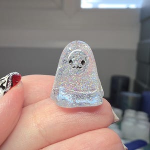 Op de afbeelding: Een klein, doorschijnend, koepelvormig spookfiguurtje gemaakt van heldere hars gevuld met fijne, veelkleurige iriserende glitters. Het spookje heeft twee eenvoudige zwarte stippen als ogen en een klein, golvend zwart lijntje als mond op de voorzijde.