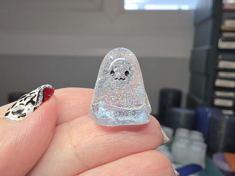 Op de afbeelding: Een klein, doorschijnend, koepelvormig spookfiguurtje gemaakt van heldere hars gevuld met fijne, veelkleurige iriserende glitters. Het spookje heeft twee eenvoudige zwarte stippen als ogen en een klein, golvend zwart lijntje als mond op de voorzijde.