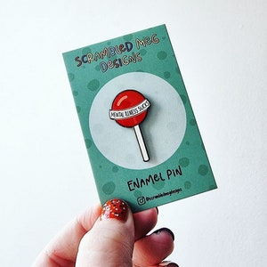 May include: A red lollipop enamel pin with the text "MENTAL ILLNESS SUCKS" on a white banner. The pin is on a teal card with the text "SCRAMBLED MEG DESIGNS" and "ENAMEL PIN".