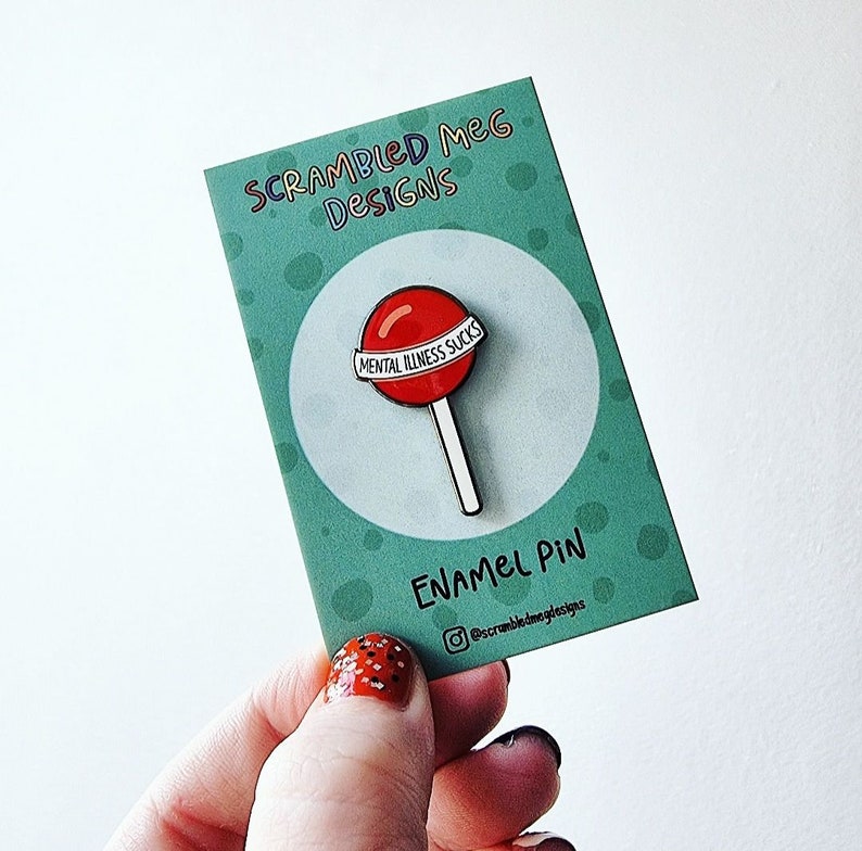 May include: A red lollipop enamel pin with the text "MENTAL ILLNESS SUCKS" on a white banner. The pin is on a teal card with the text "SCRAMBLED MEG DESIGNS" and "ENAMEL PIN".