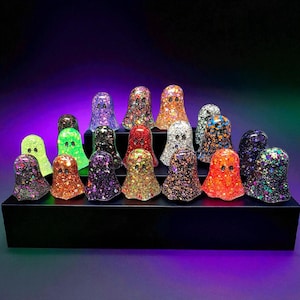 May include: A selection of glittery ghost figurines in various colours, including orange, purple, and green, arranged on a black display stand. Each ghost has two eye holes and a rounded shape, ideal for Halloween decorations.