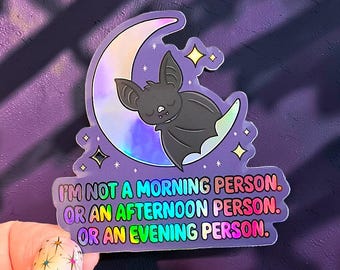 Puede incluir: Una pegatina holográfica con un murciélago gris oscuro durmiendo sobre una luna creciente. El texto dice: I'M NOT A MORNING PERSON. OR AN AFTERNOON PERSON. OR AN EVENING PERSON. Las letras tienen un degradado arcoíris sobre fondo morado.