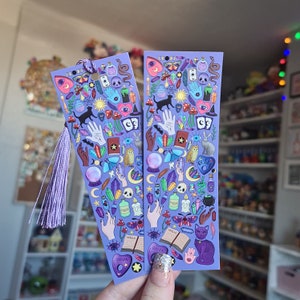May include: Two purple bookmarks with a repeating pattern of mystical symbols, including cats, hands, crystals, and potion bottles. Each bookmark has a matching purple tassel. The word "Spells" is visible on an open book.