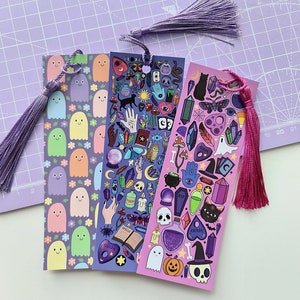 May include: Three decorative bookmarks with tassels. One features smiling ghost characters and flowers, another has mystical symbols, and the third has a Halloween theme with a witch's hat, skull, and black cat. Each bookmark has a different color tassel.