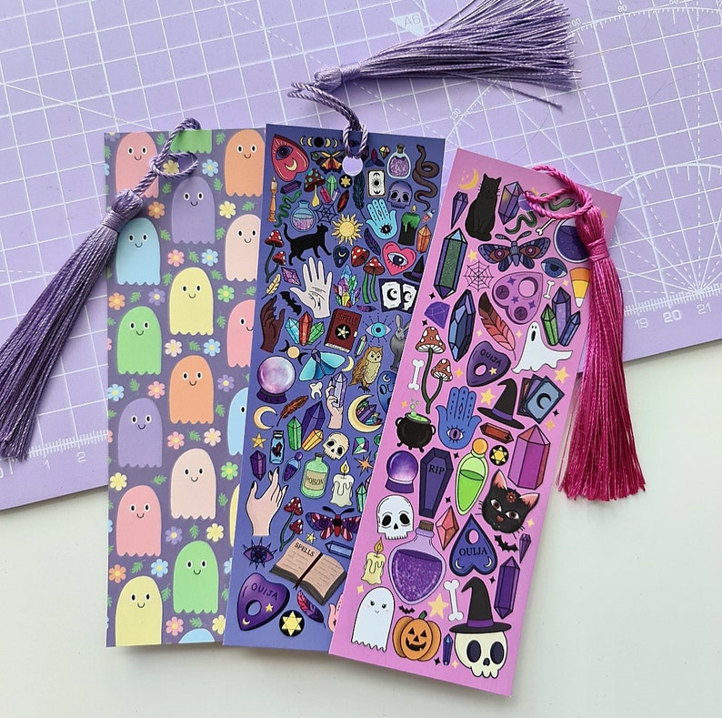 May include: Three decorative bookmarks with tassels. One features smiling ghost characters and flowers, another has mystical symbols, and the third has a Halloween theme with a witch's hat, skull, and black cat. Each bookmark has a different color tassel.