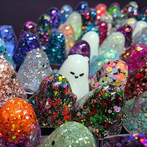 Op de afbeelding: Een verzameling kleine, spookvormige beeldjes gemaakt van hars, elk met een unieke afwerking. De figuren zijn gevuld met diverse gekleurde glitters, holografische vlokken en metallic pailletten, waarbij sommige figuren effen wit of doorschijnend zijn.
