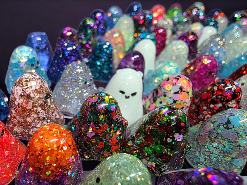 Op de afbeelding: Een verzameling kleine, spookvormige beeldjes gemaakt van hars, elk met een unieke afwerking. De figuren zijn gevuld met diverse gekleurde glitters, holografische vlokken en metallic pailletten, waarbij sommige figuren effen wit of doorschijnend zijn.