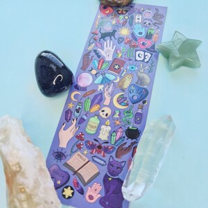 May include: A purple sticker sheet with various mystical and witchy illustrations, including a black cat, crystals, a Ouija board, and potion bottles. The sticker sheet is surrounded by crystals and a green star-shaped stone.