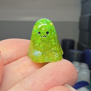 Op de afbeelding: Een klein, doorschijnend limoengroen spookfiguurtje gemaakt van hars, met ingebedde holografische glitterdeeltjes. Het spookje heeft een gladde, ronde koepelvorm met twee kleine zwarte ronde ogen en een piepklein, golvend zwart mondlijntje.