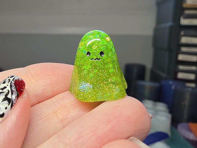 Op de afbeelding: Een klein, doorschijnend limoengroen spookfiguurtje gemaakt van hars, met ingebedde holografische glitterdeeltjes. Het spookje heeft een gladde, ronde koepelvorm met twee kleine zwarte ronde ogen en een piepklein, golvend zwart mondlijntje.
