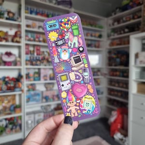 Pode incluir: Um marcador de livros roxo com ilustra&ccedil;&otilde;es coloridas de &iacute;cones da cultura pop dos anos 90, incluindo um Cubo de Rubik, Furby, Game Boy e uma boneca Troll. O marcador de livros &eacute; retangular e tem um acabamento brilhante.