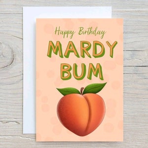 May include: A greeting card with a peach-colored background and the words "Happy Birthday MARDY BUM" in green and orange. A cartoon peach with green leaves is at the bottom. The card is on a white envelope.