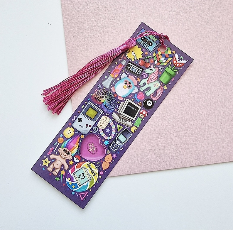Pode incluir: Marcador de livros roxo com borla rosa, apresentando ilustra&ccedil;&otilde;es coloridas de &iacute;cones dos anos 90, como um Furby, Game Boy, Cubo de Rubik e um Tamagotchi. O marcador &eacute; retangular e repousa sobre uma superf&iacute;cie rosa.