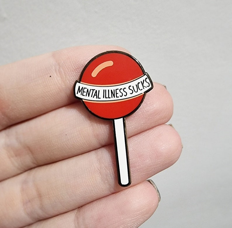 May include: A red lollipop-shaped enamel pin with a white banner that reads "MENTAL ILLNESS SUCKS." The pin has a black outline and a white stick. The lollipop has a curved orange accent.