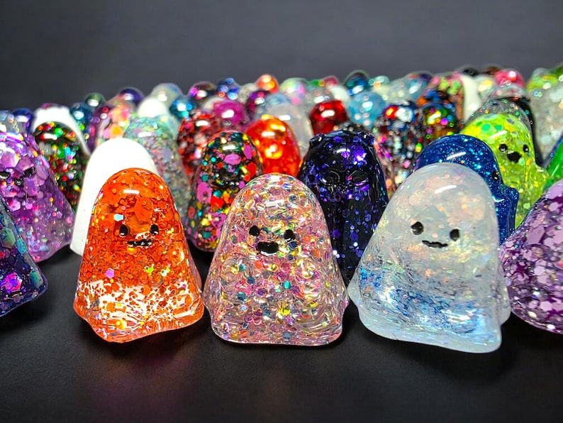 Op de afbeelding: Een verzameling kleine, doorschijnende spookvormige beeldjes gemaakt van hars. Elk spookje heeft een unieke kleur, waaronder oranje, roze, blauw, paars en wit, en is gevuld met verschillende maten iriserende en metallic glitters.