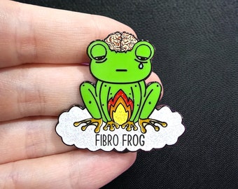 May include: A green enamel pin featuring a cartoon frog with a brain exposed, a tear, and flames. The frog sits on a cloud with the text "FIBRO FROG" in white. The pin is held by a hand.