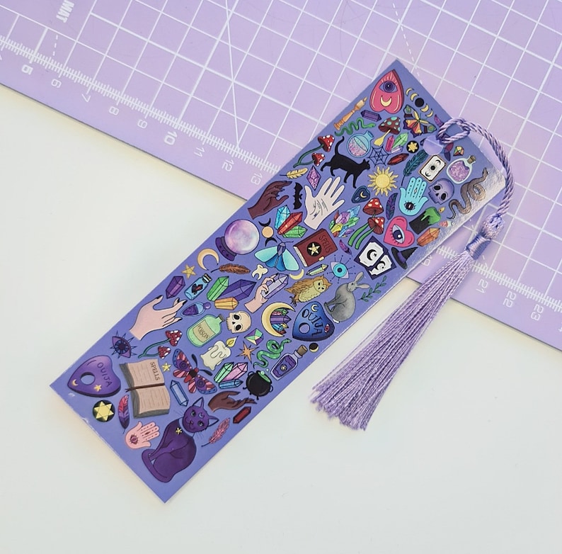 May include: A purple bookmark with a tassel, featuring illustrations of crystals, a black cat, a Ouija board, and other mystical symbols. The bookmark has the word "SPELLS" on it.