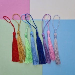 May include: A collection of colorful tassels in shades of red, gold, green, blue, purple, pink, and silver. Each tassel has a looped cord for hanging. The tassels are arranged against a backdrop of pastel colors.