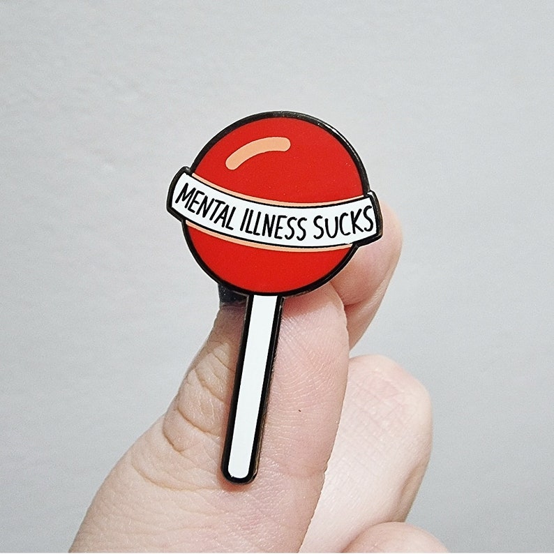 May include: A red lollipop enamel pin with a white and black stick. The pin has a white banner with the words "MENTAL ILLNESS SUCKS" in black lettering. The lollipop has a black outline.