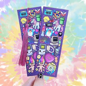 Pode incluir: Dois marcadores de livros roxos com tema retr&ocirc; dos anos 90, com ilustra&ccedil;&otilde;es coloridas de um Cubo de Rubik, Furby, unic&oacute;rnios e um Game Boy. Cada marcador de livros tem um borla rosa. O fundo &eacute; um padr&atilde;o tie-dye.