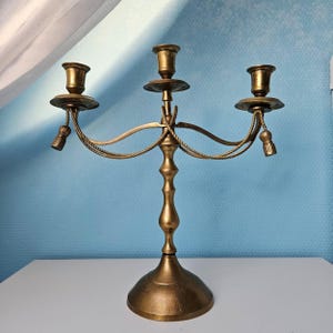 Op de afbeelding: Een messing kandelaar met drie kaarshouders. De kandelaar heeft een decoratief touwontwerp en kwastjes. De basis is rond en het ontwerp is vintage.